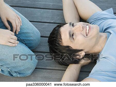Young man reclining on the ground with hands behind head, smiling, someone kneeling beside him, partial view View Large Photo Image Stock Image - Young man reclining on the ground with hands behind head, smiling, someone kneeling beside him, partial view. Fotosearch