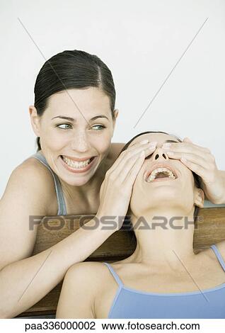 Stock Image - Young woman covering her friend's eyes, both smiling, portrait. Fotosearch
