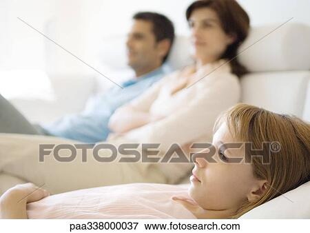 Girl reclining on sofa, parents in background View Large Photo Image Stock Photo - Girl reclining on sofa, parents in background. Fotosearch