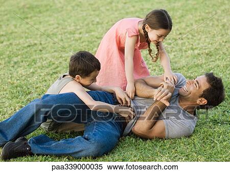 Stock Photography - Boy and girl tickling father on grass. Fotosearch