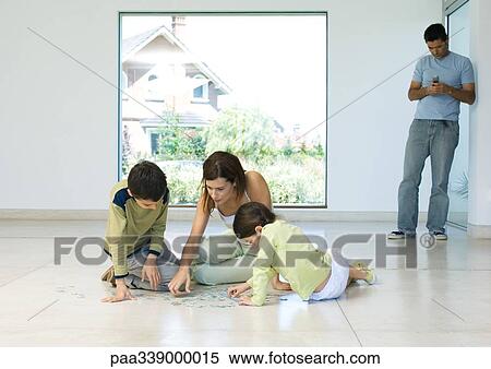 Stock Photography - Mother putting together puzzle with children while father uses cell phone in background. Fotosearch