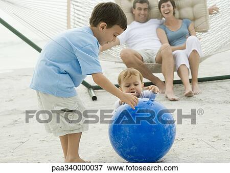 Stock Photo - Boy and baby playing with ball while parents watch from hammock. Fotosearch