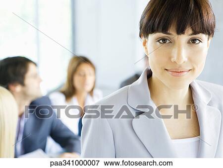 Stock Photo - Businesswoman smiling at camera, meeting in background, portrait. Fotosearch