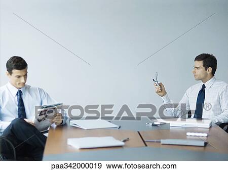 Business colleagues sitting at table, one reading magazine while other checks messages on cell phone View Large Photo Image Stock Photo - Business colleagues sitting at table, one reading magazine while other checks messages on cell phone. Fotosearch