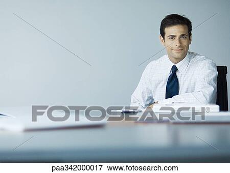 Businessman sitting at desk, smiling at camera View Large Photo Image Stock Photo - Businessman sitting at desk, smiling at camera. Fotosearch