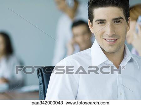 Businessman smiling at camera, colleagues in background, portrait View Large Photo Image Stock Image - Businessman smiling at camera, colleagues in background, portrait. Fotosearch