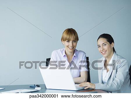 Two businesswomen using laptop, smiling at camera View Large Photo Image Stock Image - Two businesswomen using laptop, smiling at camera. Fotosearch