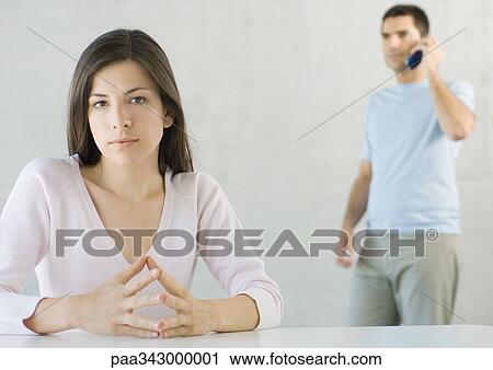 Stock Image - Woman sitting, looking at camera while man uses cell phone in background. Fotosearch