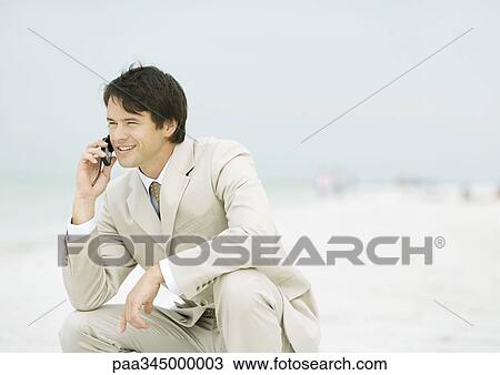 Stock Image - Businessman crouching on beach, using cell phone. Fotosearch