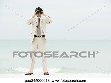 Stock Image - Businessman looking through binoculars, facing camera. Fotosearch