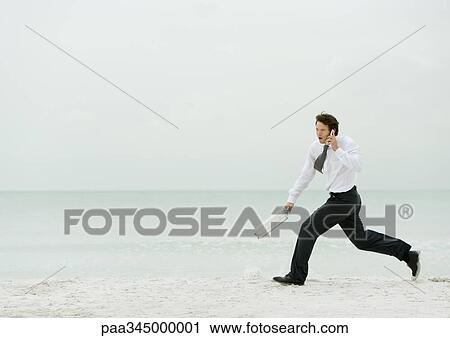 Stock Image - Businessman running on beach, with cell phone and briefcase. Fotosearch
