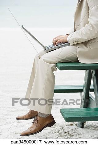 Stock Photo - Businessman sitting on picnic table on beach, using laptop, chest down. Fotosearch