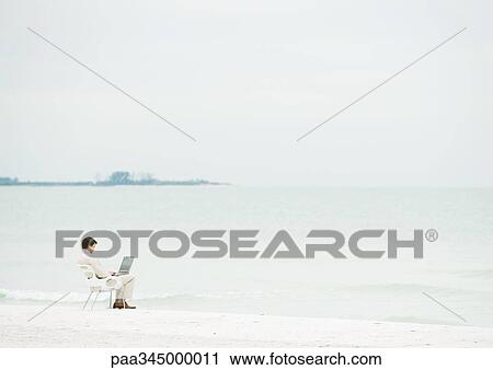 Stock Image - Businessman using laptop on beach, mid-distance. Fotosearch