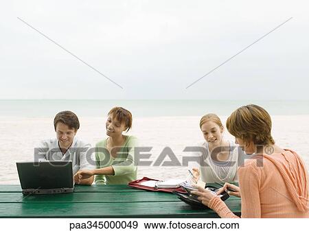 Stock Photo - Four adults sitting at picnic table on beach, looking at laptop and agendas. Fotosearch