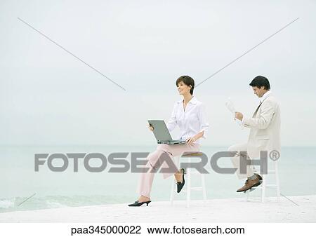 Stock Image - Two business people sitting on stools on beach, one using laptop, the other reading newspaper. Fotosearch