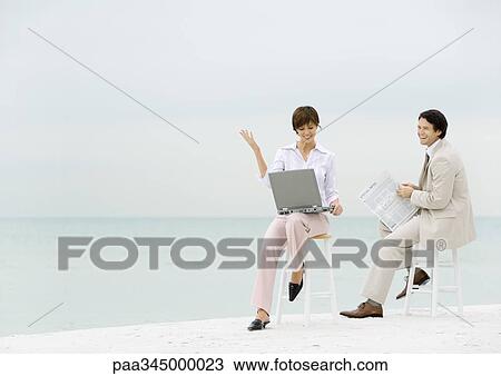 Two business people sitting on stools on beach, one using laptop, the other reading newspaper View Large Photo Image Stock Image - Two business people sitting on stools on beach, one using laptop, the other reading newspaper. Fotosearch