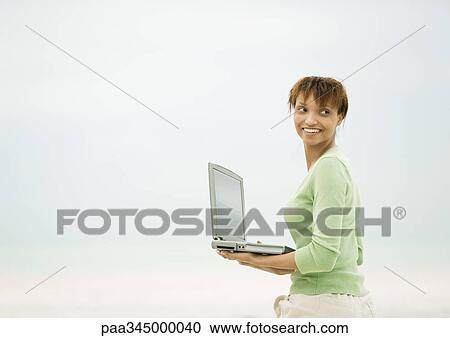Stock Image - Woman holding open laptop, looking over shoulder. Fotosearch