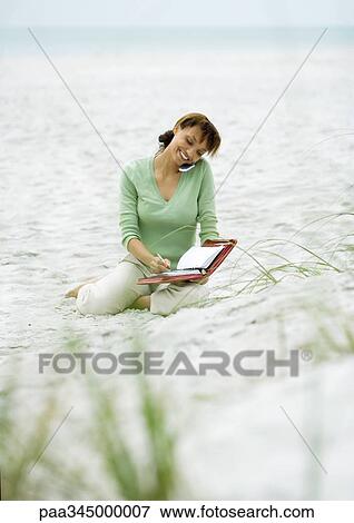 Woman sitting on sand, using cell phone and writing in agenda View Large Photo Image Stock Photo - Woman sitting on sand, using cell phone and writing in agenda. Fotosearch