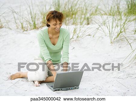 Stock Image - Woman sitting on sand, using laptop. Fotosearch