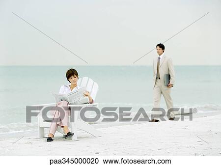 Stock Image - Woman sitting reading newspaper on beach as businessman carrying laptop approaches. Fotosearch