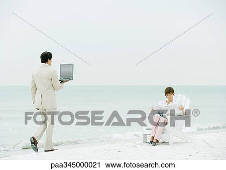 Stock Image - Woman sitting reading newspaper on beach as businessman carrying open laptop approaches. Fotosearch