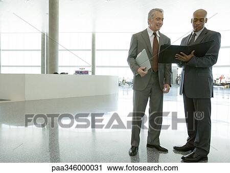 Two businessmen standing in airport concourse, looking at agenda View Large Photo Image Stock Image - Two businessmen standing in airport concourse, looking at agenda. Fotosearch