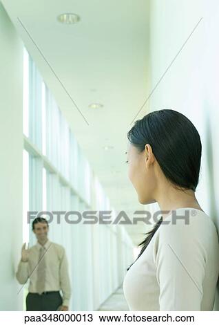 Stock Image - Woman's male colleague waving in hallway. Fotosearch