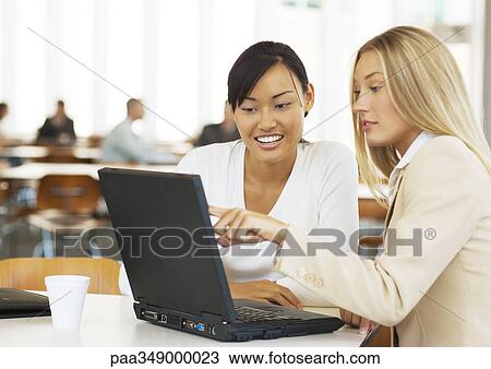 Stock Image - Two young business women using laptop together, smiling. Fotosearch