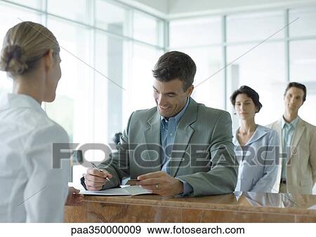 Man signing at reception desk while people wait in line behind him View Large Photo Image Stock Photo - Man signing at reception desk while people wait in line behind him. Fotosearch