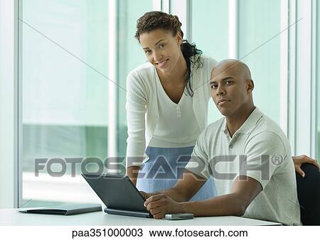 Stock Image - Businessman sitting at desk while woman leans over shoulder, smiling at camera. Fotosearch