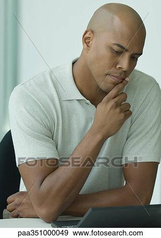 Man sitting at desk, looking at laptop and furrowing brow View Large Photo Image Stock Photo - Man sitting at desk, looking at laptop and furrowing brow. Fotosearch