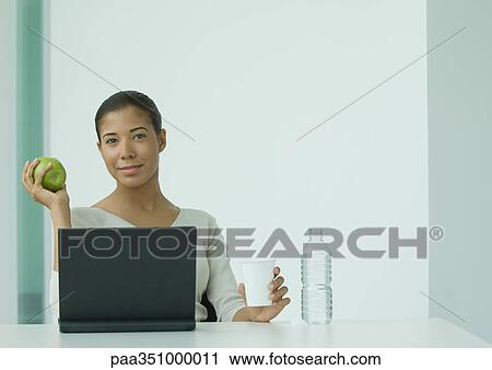 Woman sitting at laptop, holding glass of water and apple View Large Photo Image Stock Image - Woman sitting at laptop, holding glass of water and apple. Fotosearch