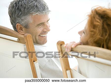 Stock Image - Mature couple, sitting in deck chairs, smiling at each other . Fotosearch
