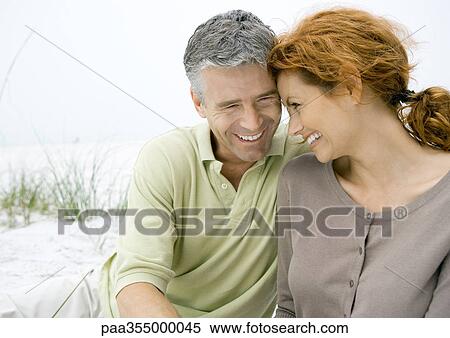 Stock Photography - Mature couple sitting on beach, smiling with heads together. Fotosearch