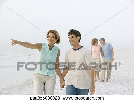 Stock Image - Mature couple walking on beach, woman pointing to distance. Fotosearch