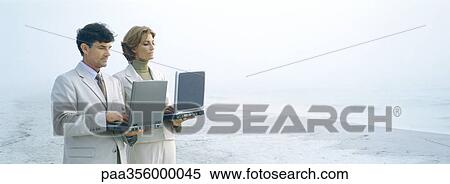 Businessman and woman standing, holding laptops on beach View Large Photo Image Stock Photography - Businessman and woman standing, holding laptops on beach. Fotosearch