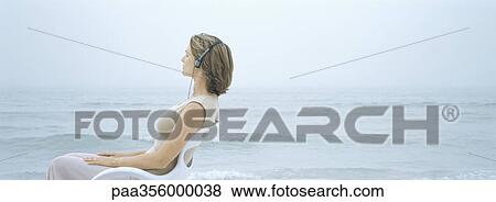 Woman sitting in chair, listening to headphones, sea in background View Large Photo Image Stock Photo - Woman sitting in chair, listening to headphones, sea in background. Fotosearch