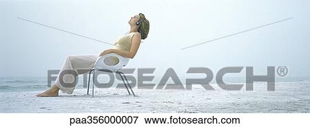 Stock Photo - Woman sitting in chair on beach, wearing headphones. Fotosearch