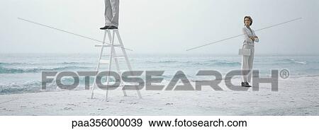 Woman standing on beach with arms crossed, looking toward man standing on ladder View Large Photo Image Stock Photo - Woman standing on beach with arms crossed, looking toward man standing on ladder. Fotosearch