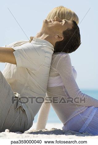Stock Image - Couple sitting back to back on beach. Fotosearch
