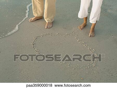 Stock Photo - Couple standing by heart drawn in sand, view of knee down. Fotosearch