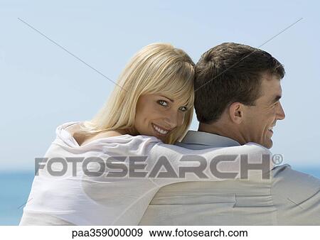 Stock Photo - Woman leaning against man, looking over shoulder at camera, sea in background. Fotosearch
