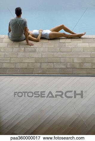 Stock Photo - Couple by edge of pool, woman lying down and man sitting. Fotosearch
