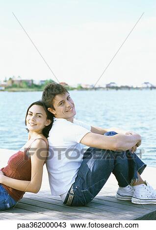 Stock Image - Young couple sitting back to back on dock. Fotosearch