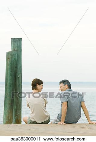 Stock Image - Couple sitting on dock, talking, rear view. Fotosearch