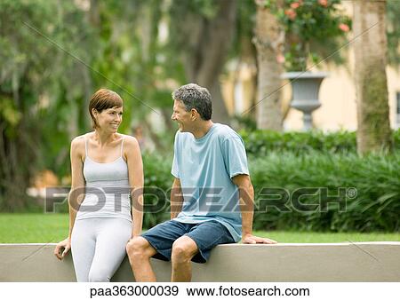 Stock Photo - Mature man and woman in workout clothes sitting on low wall, talking. Fotosearch