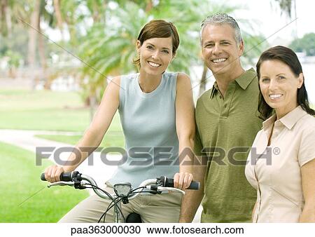 Three adult friends smiling at camera, one on bike View Large Photo Image Stock Image - Three adult friends smiling at camera, one on bike. Fotosearch