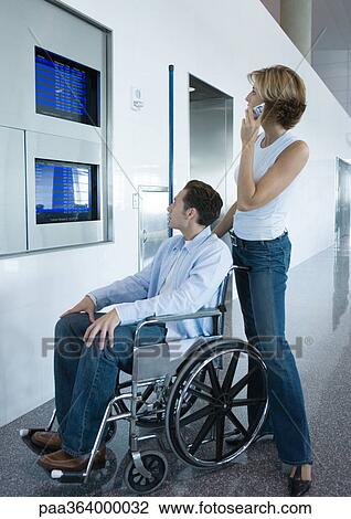 Stock Image - Woman and man in wheelchair looking at departure board in airport. Fotosearch