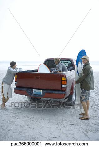Stock Image - Young adults standing around pick-up truck on beach. Fotosearch