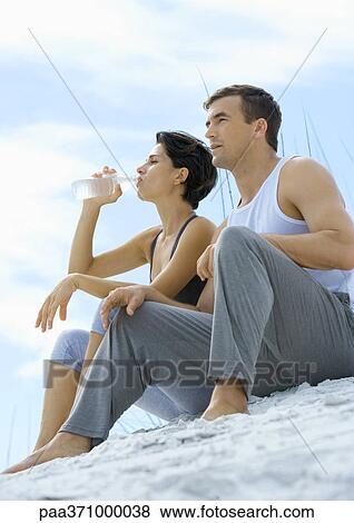 Couple sitting in exercise clothing on beach, woman drinking water View Large Photo Image Stock Photo - Couple sitting in exercise clothing on beach, woman drinking water. Fotosearch
